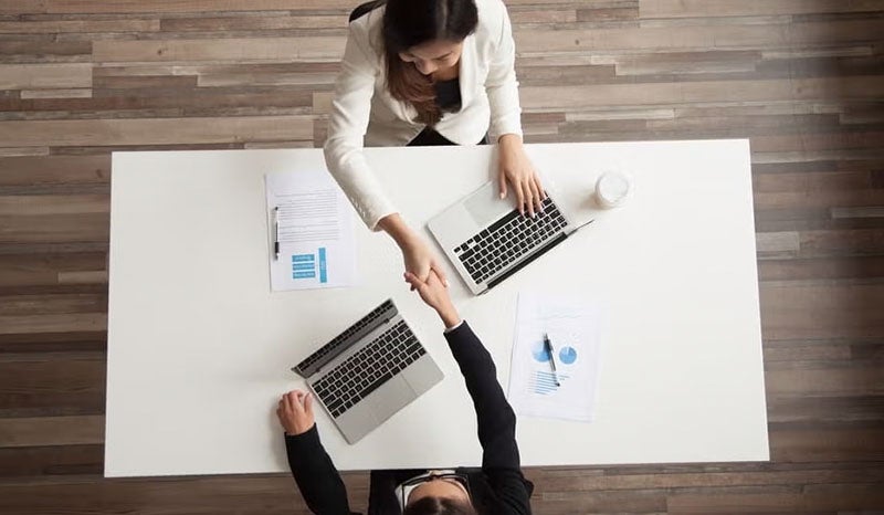 Two professionals shaking hands in an office, with documents and laptops on the table.