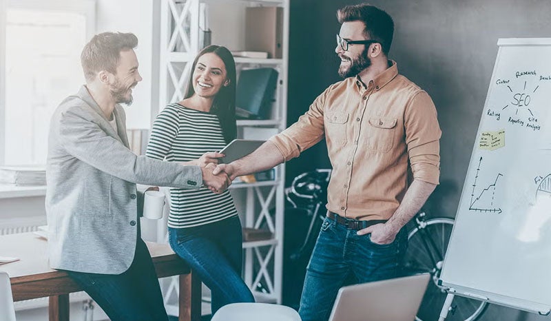 A team of three people shaking hands and smiling in a collaborative office setting.