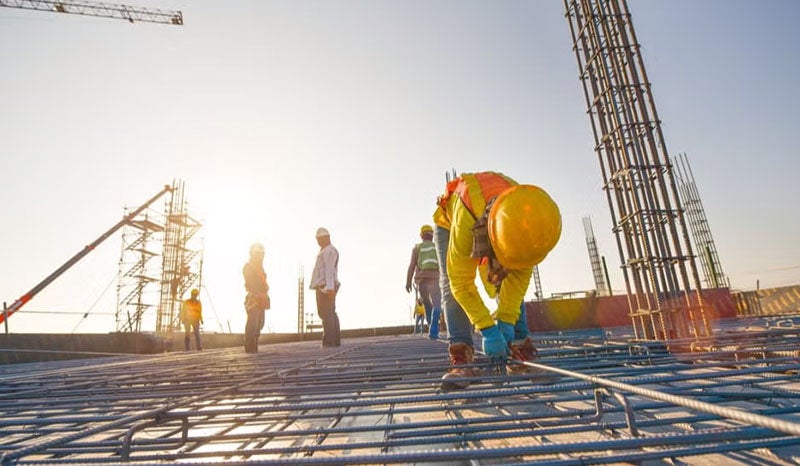 Construction workers on site, with one man working on a steel frame under the sun.