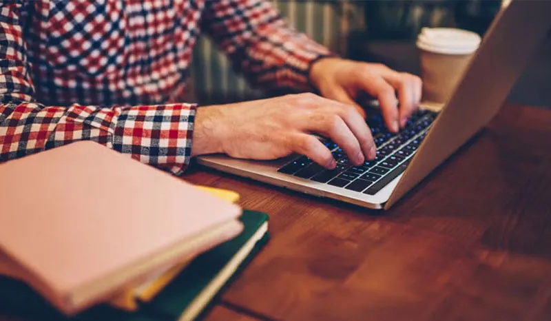 A person typing on a laptop in a cozy workspace with books and coffee.