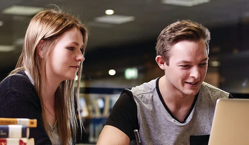 Two students collaborating and working on a laptop in a library setting.