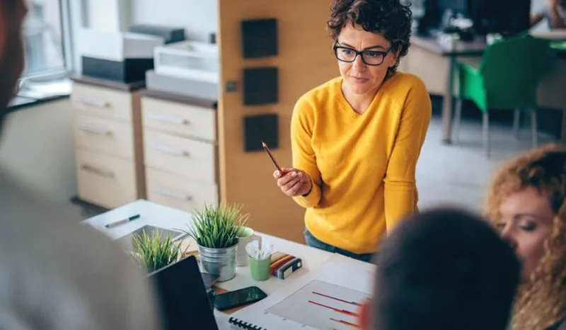 A woman in a yellow shirt leading a discussion with colleagues in an office.
